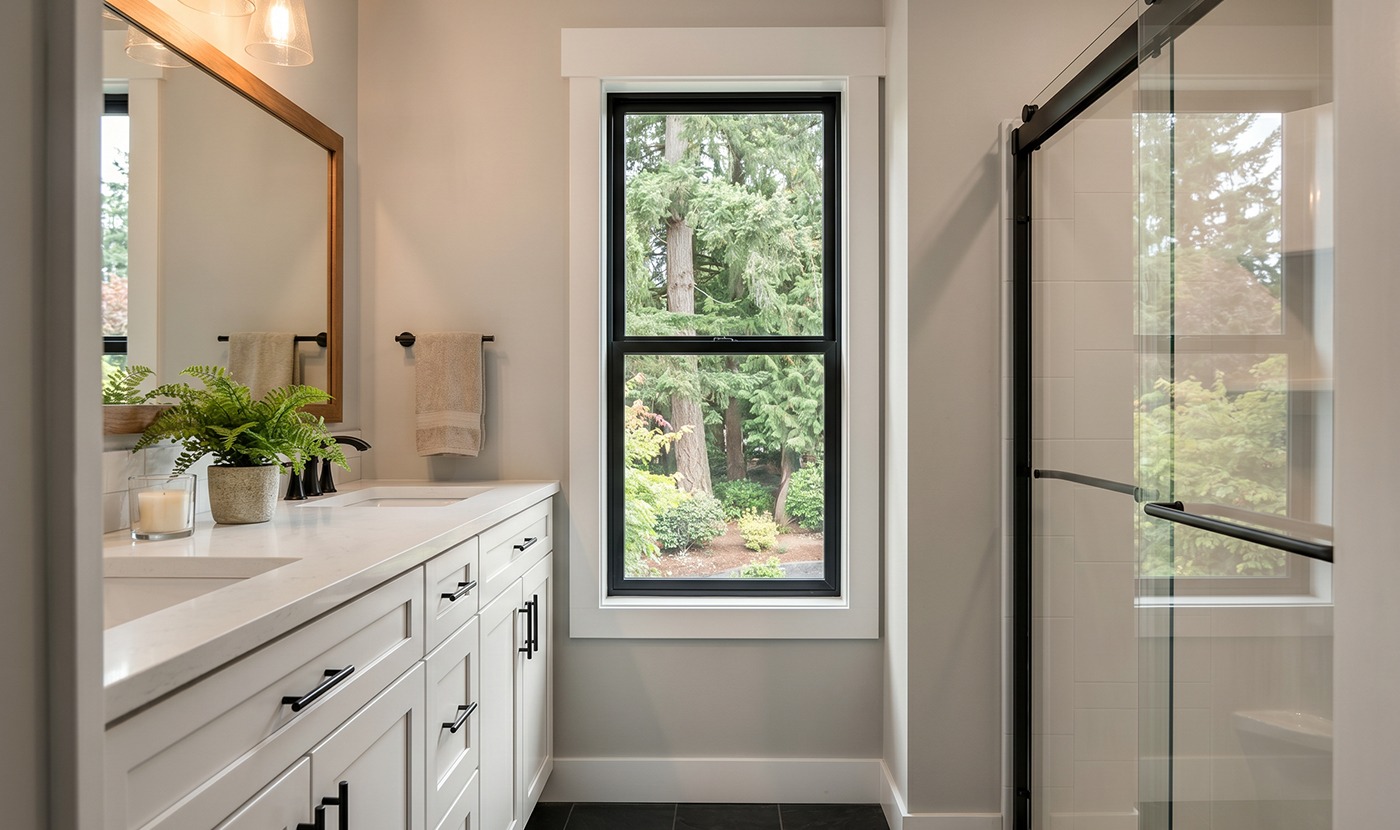 Bright modern bathroom with white cabinetry, marble countertop, and a window overlooking greenery by a glass shower enclosure.