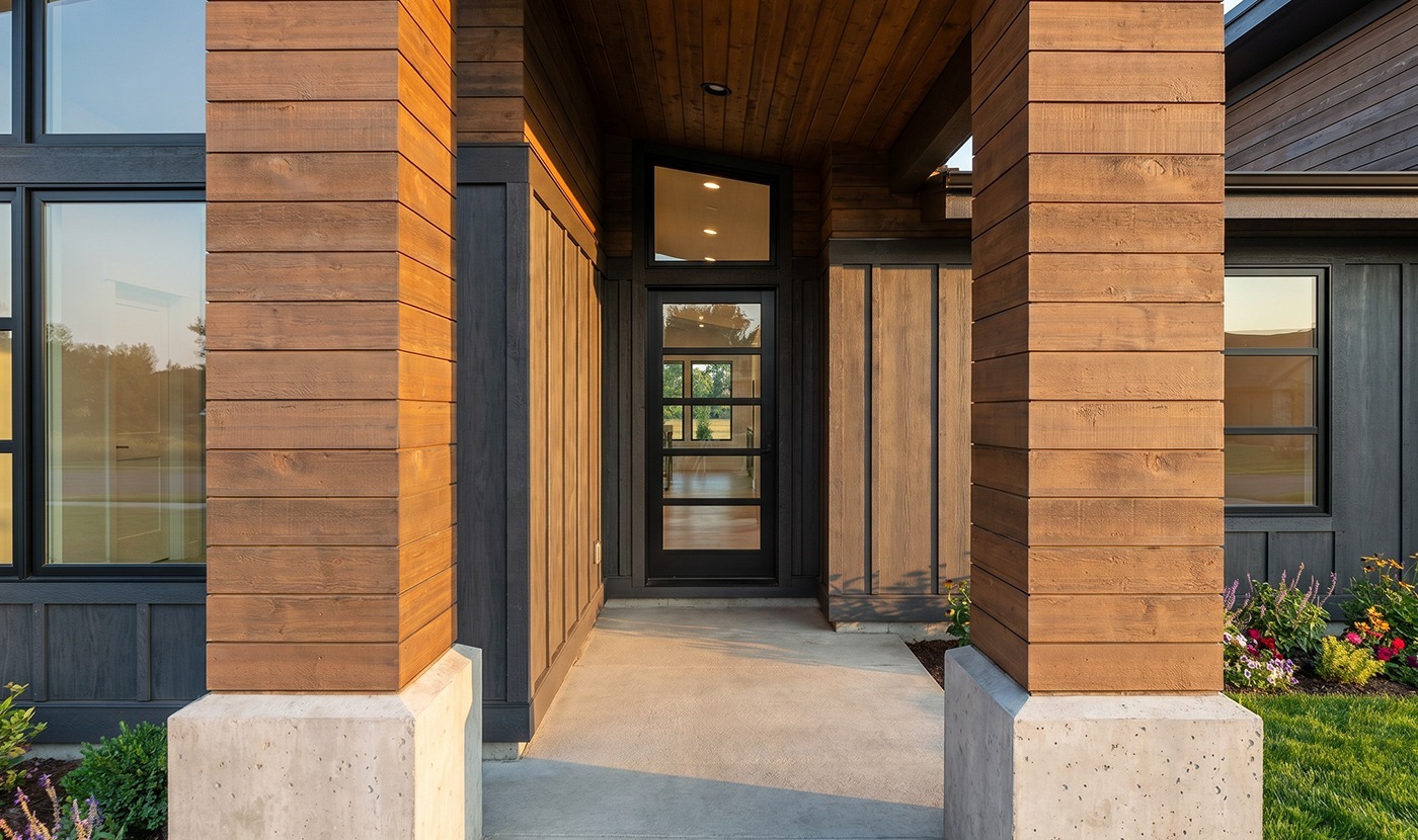 Modern house entrance with two wooden columns, glass front door, and concrete porch, warm lighting inside cools the entryway glow.