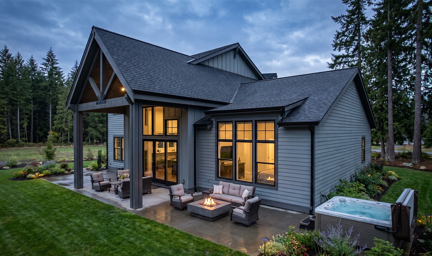 Modern gray house with a covered patio, warm outdoor seating, and a glowing fire pit at dusk.