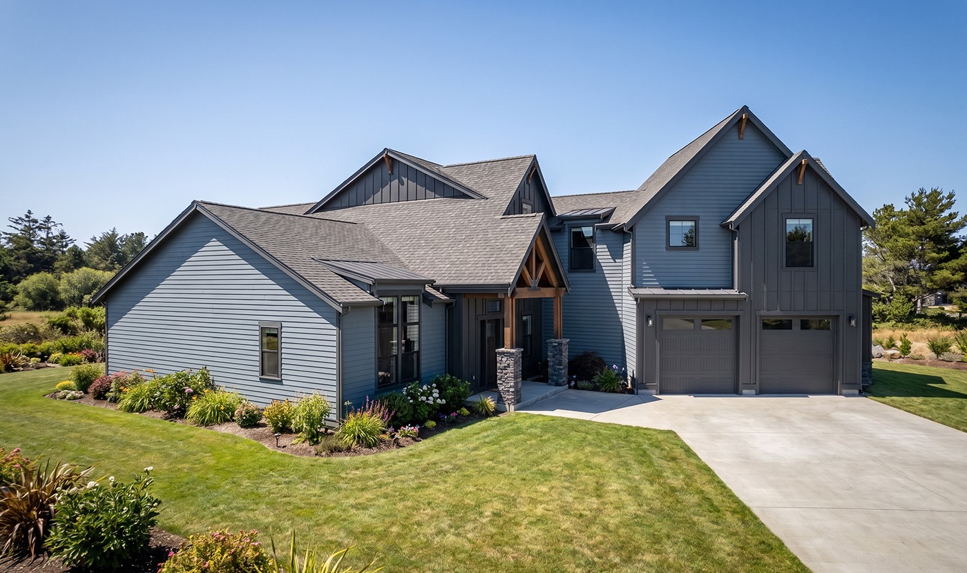 Modern gray two-story house with a three-car garage, stone pillars, and a landscaped front yard under a clear blue sky.