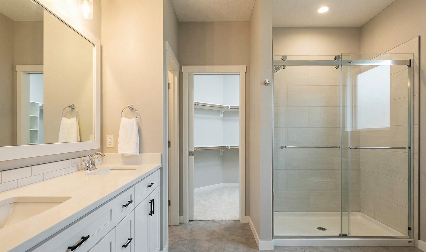 Bright modern bathroom with double-sink vanity, glass-enclosed shower, and a walk-in closet visible through a doorway.
