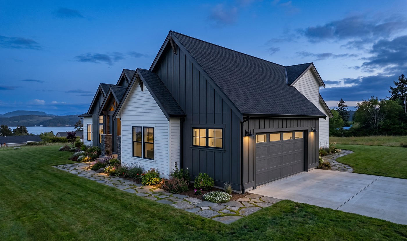Modern house with dark vertical siding and a two-car garage, lit windows, stone pathway, and a manicured lawn at dusk.