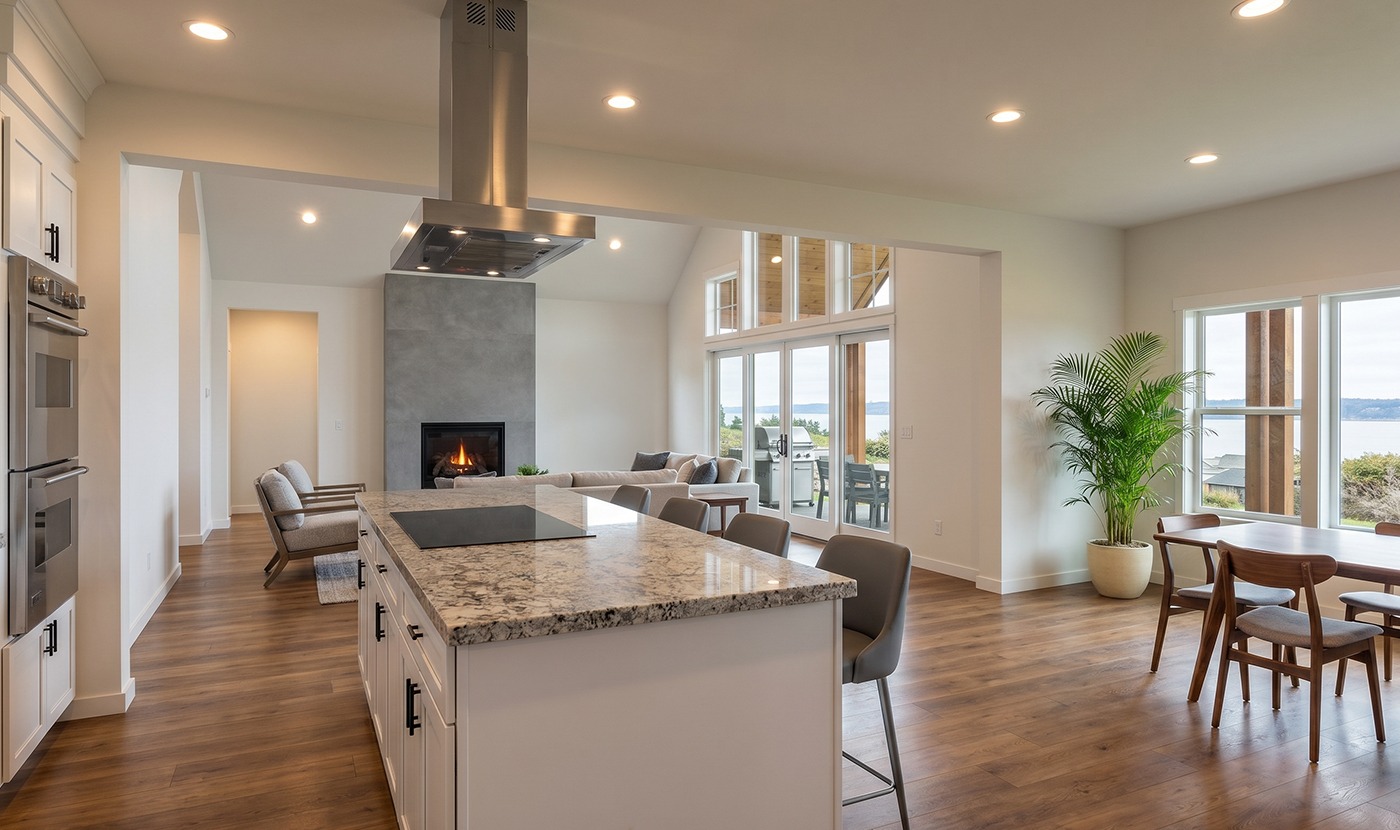 Open-concept kitchen and living area with marble island, stainless range hood, fireplace, and large windows showing an outdoor view.