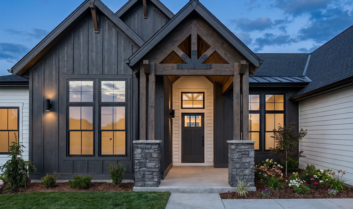Front entrance of a modern craftsman-style home with dark wood siding, stone columns, and warm interior lighting visible through large windows